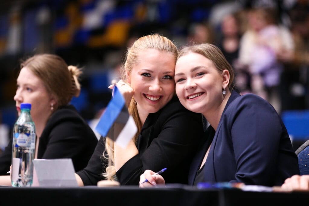 Kohtunikud ja korraldajad võimlemisvõistlusel naeratamas žüriilaua taga – toetav meeleolu saalis.